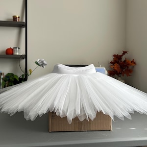 May include: A white ballet tutu with a wide, layered skirt. The tutu is made of sheer fabric and has a fitted waistband. The skirt is full and flared, creating a classic ballet silhouette. The tutu is displayed on a cardboard box.