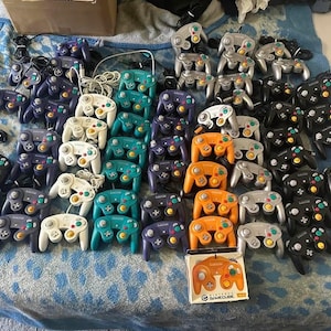 Könnte beinhalten: Eine Sammlung verschiedenfarbiger Nintendo GameCube-Controller, darunter schwarz, lila, silber, orange, weiß und türkis. Einige Controller befinden sich noch in der Originalverpackung. Die Controller sind auf einer blau gemusterten Oberfläche angeordnet.