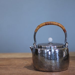 Könnte beinhalten: Eine silberne Teekanne mit gehämmerter Oberfläche und einem geflochtenen braunen Griff. Die Teekanne hat einen weißen Keramikknopf auf dem Deckel.
