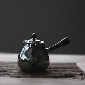 Pode incluir: Uma chaleira de prata com uma alça preta e um botão preto na tampa. A chaleira está sobre uma superfície de madeira.