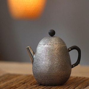 May include: A silver teapot with a textured surface and a black knob on the lid. The teapot has a spout and a handle.