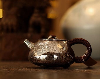 Puede incluir: Una pequeña tetera de metal martillado con un asa tejida marrón y detalles en la tapa. La tetera tiene un pico plateado y una superficie texturizada. El fondo está borroso, con tonos cálidos.