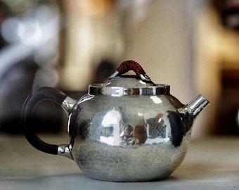 Puede incluir: Una tetera de plata con un acabado martillado y un asa de cuero marrón. La tetera tiene un cuerpo redondo y un pico en el lateral.