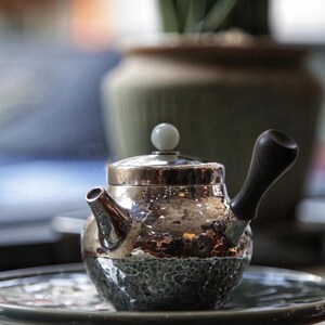 May include: A silver teapot with a hammered finish and a jade green knob on the lid. The teapot has a dark wood handle and is sitting on a gray and white patterned surface.