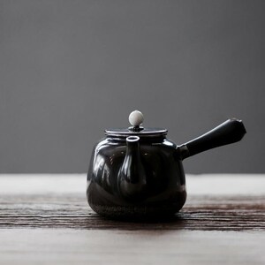 May include: A black ceramic teapot with a white knob on the lid and a black wooden handle. The teapot is sitting on a wooden surface.