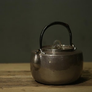 Pode incluir: Uma chaleira de prata com uma alça preta e uma tampa de prata. A chaleira tem um acabamento martelado e está sobre uma superfície de madeira.