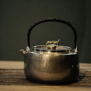 May include: A silver teapot with a hammered finish and a black leather handle. The lid has a gold-colored, twisted wire knob.