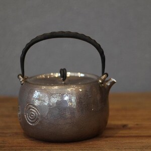 May include: A silver teapot with a hammered finish and a black handle. The teapot has a round body and a spout on the side. The teapot is decorated with a swirling pattern.
