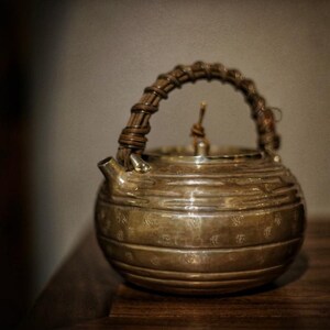 May include: A silver teapot with a woven bamboo handle. The teapot has a rounded body with a textured surface and a decorative design.