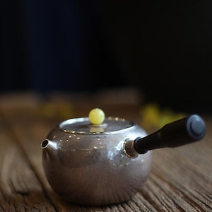 Pode incluir: Uma chaleira de prata com acabamento martelado e uma alça de madeira. A chaleira tem um botão amarelo na tampa.