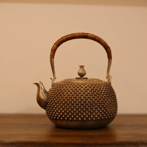 May include: A silver teapot with a brown woven handle and a textured, dimpled surface. The teapot has a small, rounded spout and a knob on the lid shaped like a flower bud.