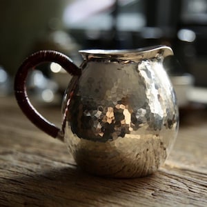 May include: A hammered silver creamer with a dark brown wrapped handle. The creamer has a spout and sits on a wooden surface. The silver surface reflects the light, creating a textured appearance. The creamer is likely used for serving cream or milk.