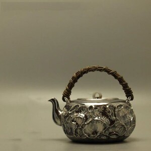 Pode incluir: Uma chaleira de prata com uma alça trançada marrom e um design floral. A chaleira tem uma tampa com uma pequena pega.