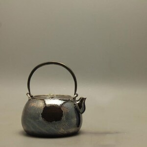 May include: A silver teapot with a hammered finish and a black handle. The teapot has a round body and a spout on the side.