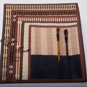 Puede incluir: Un estuche enrollable hecho de material tejido estilo bambú con ribete de tela marrón y una sección de bolsillo azul oscuro. Dos pinceles con mangos de madera y cerdas oscuras están insertados en los bolsillos de la alfombrilla organizadora.