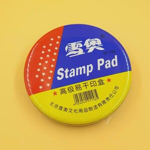 Könnte beinhalten: Ein runder Stempelkissenbehälter mit einem mehrfarbigen Deckel in den Farben Rot, Blau und Gelb. Der Deckel zeigt den Text "Stamp Pad", chinesische Schriftzeichen, einen Strichcode und ein Muster aus kleinen weißen Sternen auf dem roten Bereich.