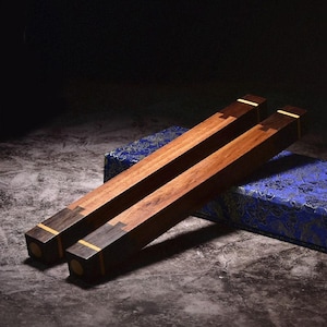 May include: A pair of long, square-profile wooden chopsticks featuring a natural light brown wood center section, dark brown wood ends, and thin yellow accent bands. The chopsticks have a visible dovetail-style joinery detail at the ends.
