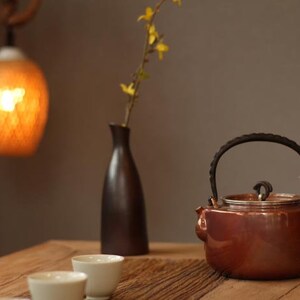May include: A brown teapot with a black handle and lid sits on a wooden table with three white teacups. A vase with yellow flowers and a hanging light fixture are in the background.