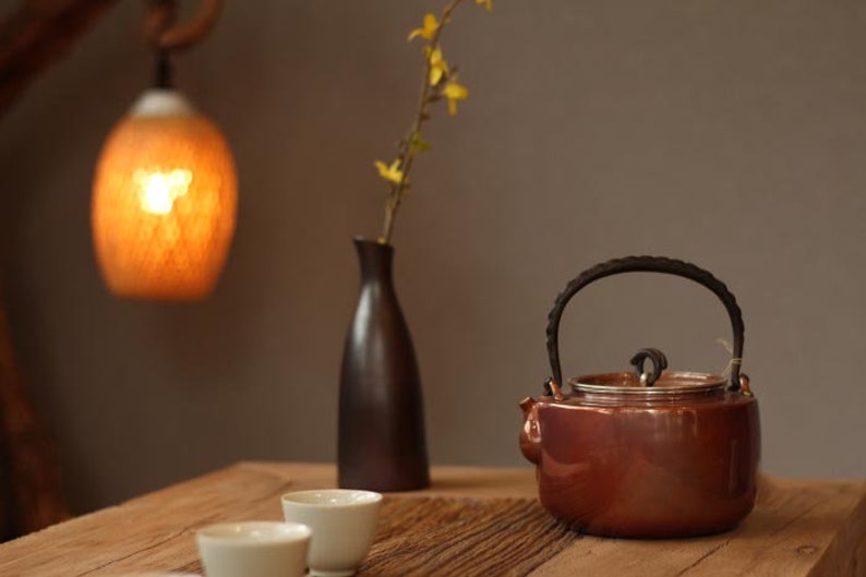 May include: A brown teapot with a black handle and lid sits on a wooden table with three white teacups. A vase with yellow flowers and a hanging light fixture are in the background.