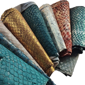 May include: A collection of stacked leather sheets featuring a textured snakeskin pattern in various metallic finishes including gold, teal, silver, copper, and dark green, showcasing a repeating scale design across the surface of each piece.