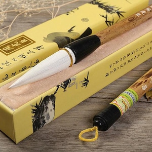 Puede incluir: Un pincel de caligrafía tradicional con un mango largo de madera marrón claro con manchas oscuras y cerdas blancas. El pincel descansa en una caja rectangular amarilla decorada con ilustraciones en tinta negra y caracteres chinos.