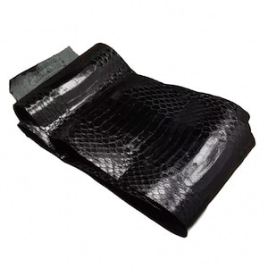 Puede incluir: Una pieza doblada de cuero de serpiente negro que presenta un patrón de escamas hexagonales en relieve con un acabado brillante y reflectante. El material está superpuesto, mostrando una parte inferior oscura y lisa en el borde superior.