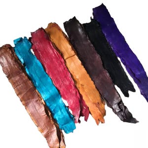 May include: A collection of seven long, narrow strips of textured fish leather arranged diagonally. The colors include brown, bright turquoise, deep red, golden yellow, dark brown, black, and vibrant purple, each showing a distinct scale pattern.