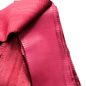 Puede incluir: Una piel de serpiente de color rosa intenso con un patrón de escamas distintivo y repetitivo en toda la superficie. El material está doblado, revelando tanto el lado exterior texturizado y estampado como el lado inferior más suave, similar al ante.