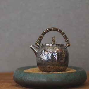 May include: A hammered metal teapot with a woven handle and a spout. The teapot sits on a teal-colored ceramic tray filled with sand. The handle is wrapped with a brown material and silver wire.
