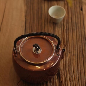 May include: A copper teapot with a black handle and a silver knob on the lid. The teapot is sitting on a wooden surface.