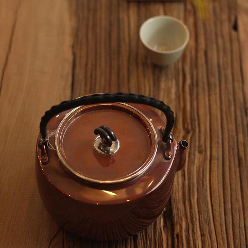 May include: A copper teapot with a black handle and a silver knob on the lid. The teapot is sitting on a wooden surface.