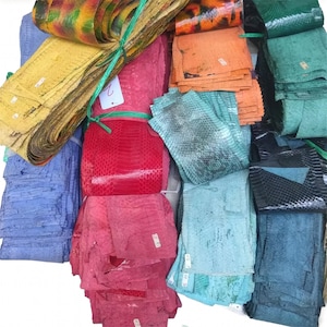 Puede incluir: Una colección de piezas rectangulares apiladas de piel de serpiente texturizada en una variedad de colores vibrantes, incluyendo amarillo, rojo, naranja, azul, verde y negro. Cada pieza presenta un patrón de escamas distinto y pequeñas etiquetas blancas.