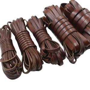 May include: Four bundles of dark brown leather cord arranged in a row. Each bundle consists of a long, flat strip of leather coiled tightly into a cylindrical shape. The leather has a smooth, matte finish and a consistent dark brown color throughout.