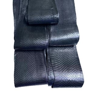 Puede incluir: Una colección de tiras largas y estrechas de cuero de piel de serpiente negra con un patrón de escamas repetitivo y distintivo. El material tiene un acabado liso y ligeramente reflectante, dispuesto en capas superpuestas.