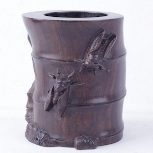 Puede incluir: Un portalápices cilíndrico de madera de color marrón oscuro con tallas en relieve detalladas de dos cigarras en la superficie exterior. El recipiente tiene un acabado liso y pulido con patrones de veta de madera visibles.