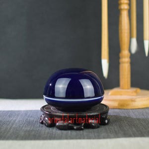Op de afbeelding: Een diepblauwe, gladde, koepelvormige keramische inktpot met een dunne witte horizontale lijn in het midden. De pot rust op een donkere, fijn uitgesneden houten voet, geplaatst voor een houten standaard voor het ophangen van penselen.