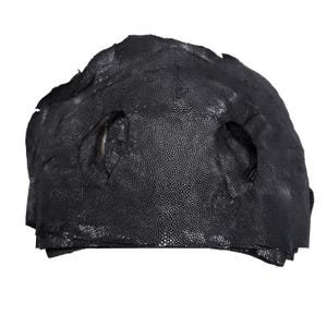 May include: A black stingray leather hide featuring a distinct pebbled texture with a dense pattern of small, raised, bead-like bumps. The hide has an irregular, organic shape with two prominent, oval-shaped openings positioned near the centre.