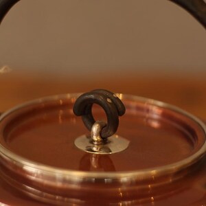 May include: Close-up of a copper kettle with a silver lid and a black iron handle. The handle is attached to the lid with a silver ring.