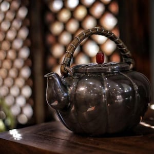 Pode incluir: Uma chaleira de prata com uma alça de madeira e um botão vermelho na tampa. A chaleira está sobre uma mesa de madeira.