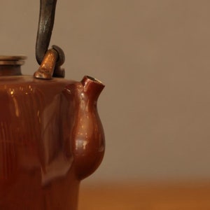 May include: A brown ceramic teapot with a metal handle and spout. The teapot has a glossy finish and a rounded shape.