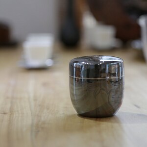 May include: A black ceramic container with a silver band and a floral design. The container is sitting on a wooden surface.
