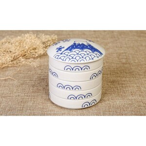 May include: A cylindrical white ceramic stackable container with a lid, featuring blue hand-painted designs of Mount Fuji, stylized waves, and cherry blossoms. The container consists of four distinct sections that fit together vertically.