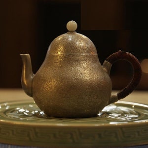 May include: A silver teapot with a pear-shaped body and a brown handle. The teapot has a textured surface and a white knob on the lid.
