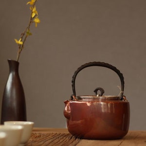 May include: A copper teapot with a black iron handle and lid. The teapot is sitting on a wooden surface.