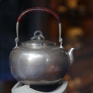 May include: A silver teapot with a hammered finish and a brown leather handle. The teapot has a round body and a spout on the side. It is sitting on a white stand.