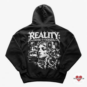 Może przedstawiać: Czarna bluza z kapturem z białą grafiką na plecach. Wzór zawiera napis REALITY DEMO VERSION, głowę klasycznego posągu, elementy niebieskie, okna komputerowe oraz dłoń wyciągniętą w stronę przestrzeni kosmicznej.