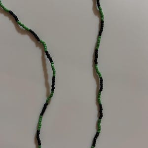 May include: Two beaded strands, one on the left and one on the right, with a repeating pattern of black and green beads. The beads are small and round, creating a textured appearance. The background is a plain white surface.