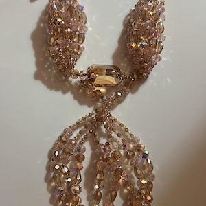 Puede incluir: Un collar glamuroso con múltiples hilos de cuentas rosas claras iridiscentes. El collar tiene grandes colgantes y detalles de cristal ámbar rectangulares y facetados. El diseño es intrincado y elegante, adecuado para ocasiones especiales.