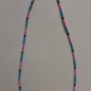 May include: A beaded necklace featuring a pattern of pink, light blue, and black beads. The necklace is made of small, round beads strung together to form a delicate chain. The beads are arranged in alternating colors, creating a visually appealing design.