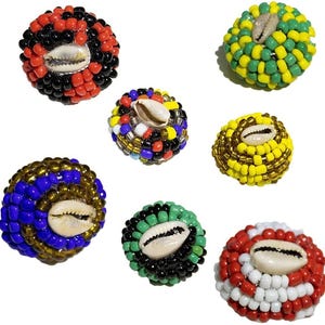 May include: A collection of seven beaded ornaments in various colors, including red, black, green, yellow, and blue. Each ornament features a cowrie shell at its center. The beads are small and tightly packed, creating a textured surface.